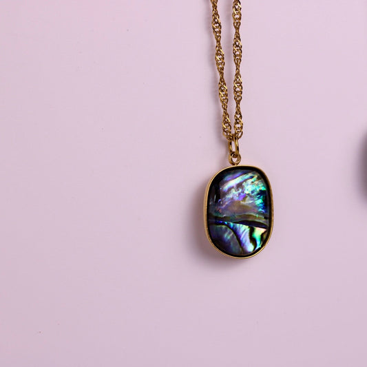 Two oval abalone shell pendant necklaces hanging against a soft pink background, one in gold-tone chain and bezel and one in silver-tone chain and bezel.