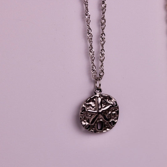 Two round pendant necklaces with Celtic-style knotwork designs, one silver-toned and one gold-toned, hanging against a solid light pink background.