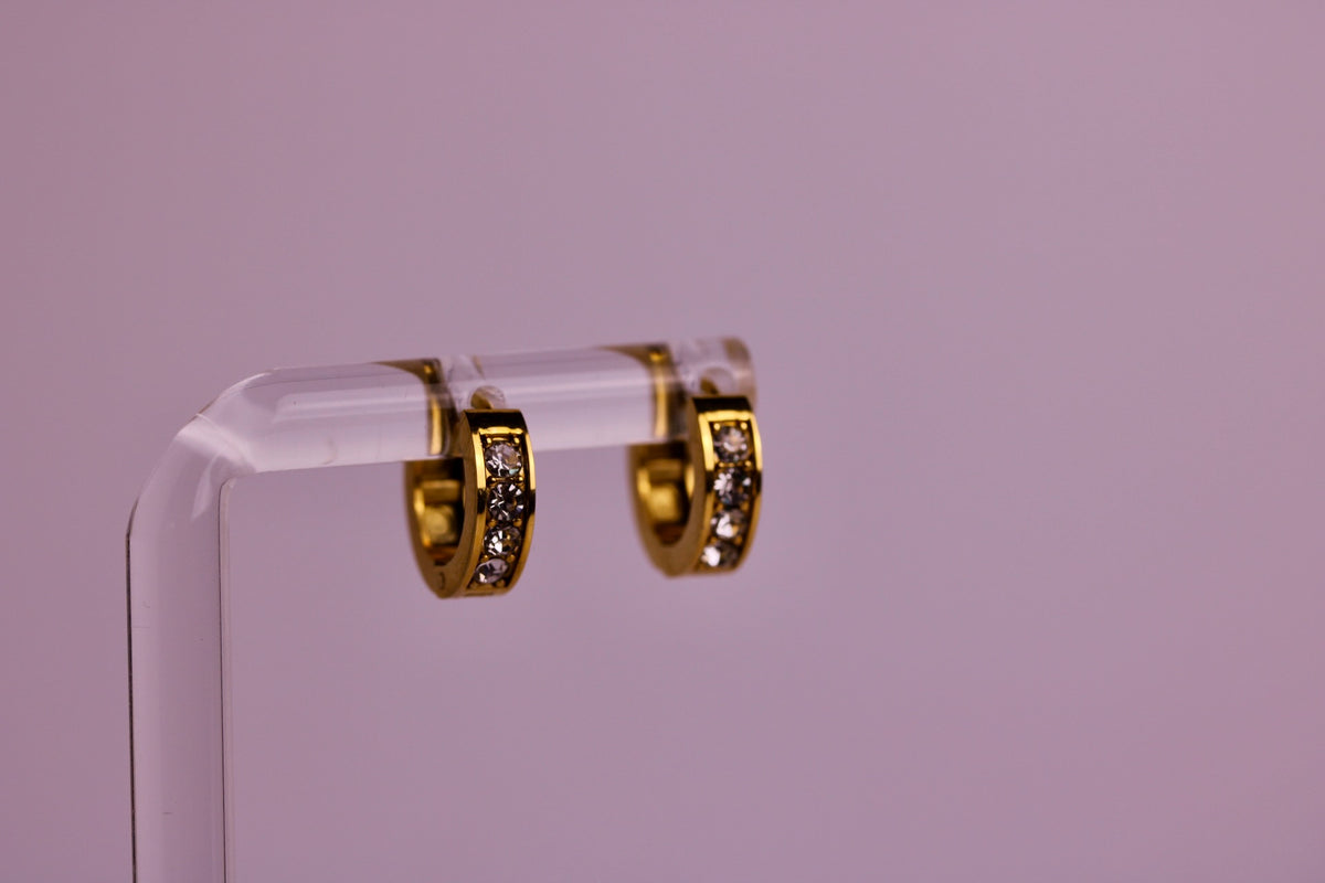 A pair of gold huggie hoop earrings with pavé-set clear stones, displayed on a clear acrylic stand against a solid lavender background.