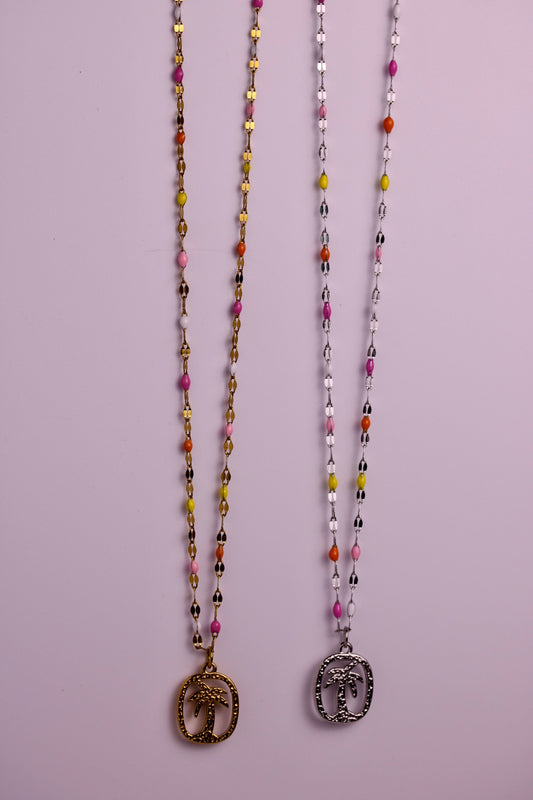 Two beaded necklaces with elephant pendants hanging against a solid light purple background. The left necklace has a gold-toned pendant and the right has a silver-toned pendant, both featuring multi-colored beads.