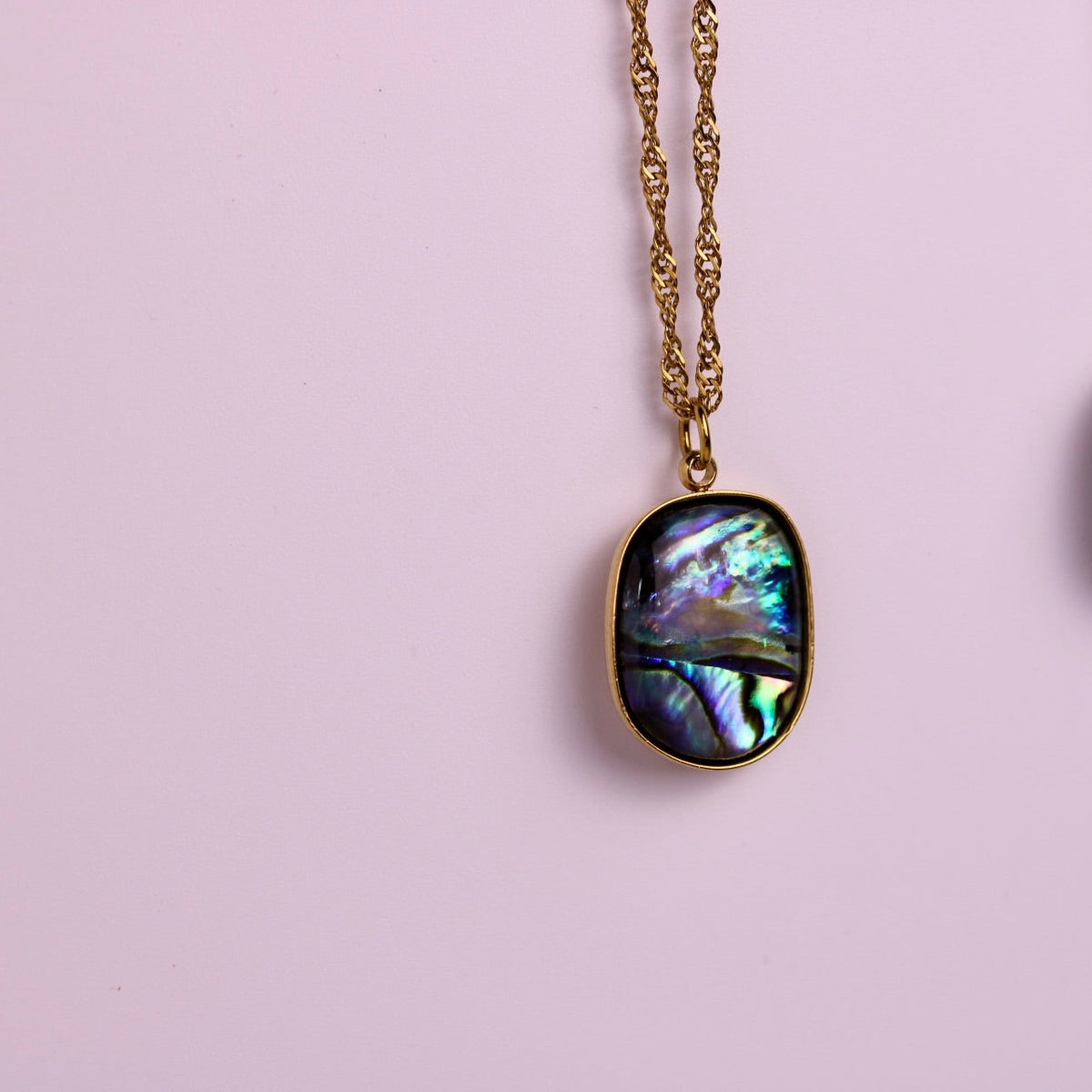 Two oval abalone shell pendant necklaces hanging against a soft pink background, one in gold-tone chain and bezel and one in silver-tone chain and bezel.