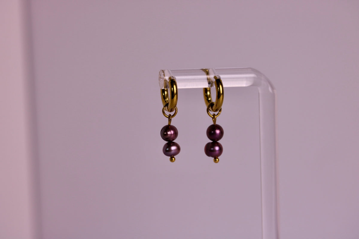 A pair of gold hoop earrings with two dark purple pearl drops, hanging from a clear acrylic display stand against a soft pink background.