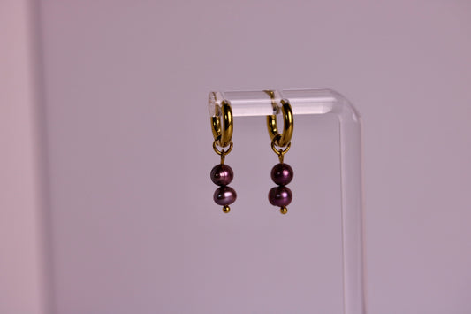 A pair of gold hoop earrings with two dark purple pearl drops, hanging from a clear acrylic display stand against a soft pink background.