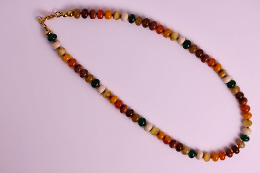 A beaded necklace made of small polished round beads in earthy brown, orange, cream, and dark green, with a small gold clasp, laid on a light pink background.