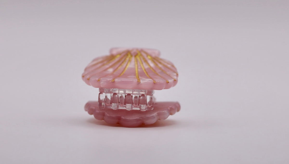 A small pink clam-shell-shaped hair claw clip with gold metallic rays on the top shell and a clear plastic inner mechanism, shown on a plain light gray background.