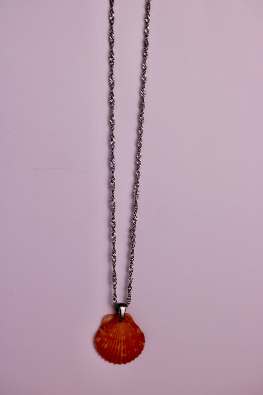 A silver-toned chain necklace with a textured orange-red scallop shell pendant, displayed against a solid light pink background.