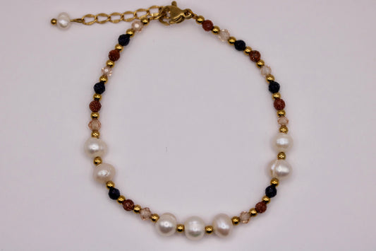 A beaded necklace featuring large white freshwater pearls, small gold spacer beads, dark black beads, and translucent brown beads, with a gold-tone lobster clasp and extender chain, displayed on a light off-white background.