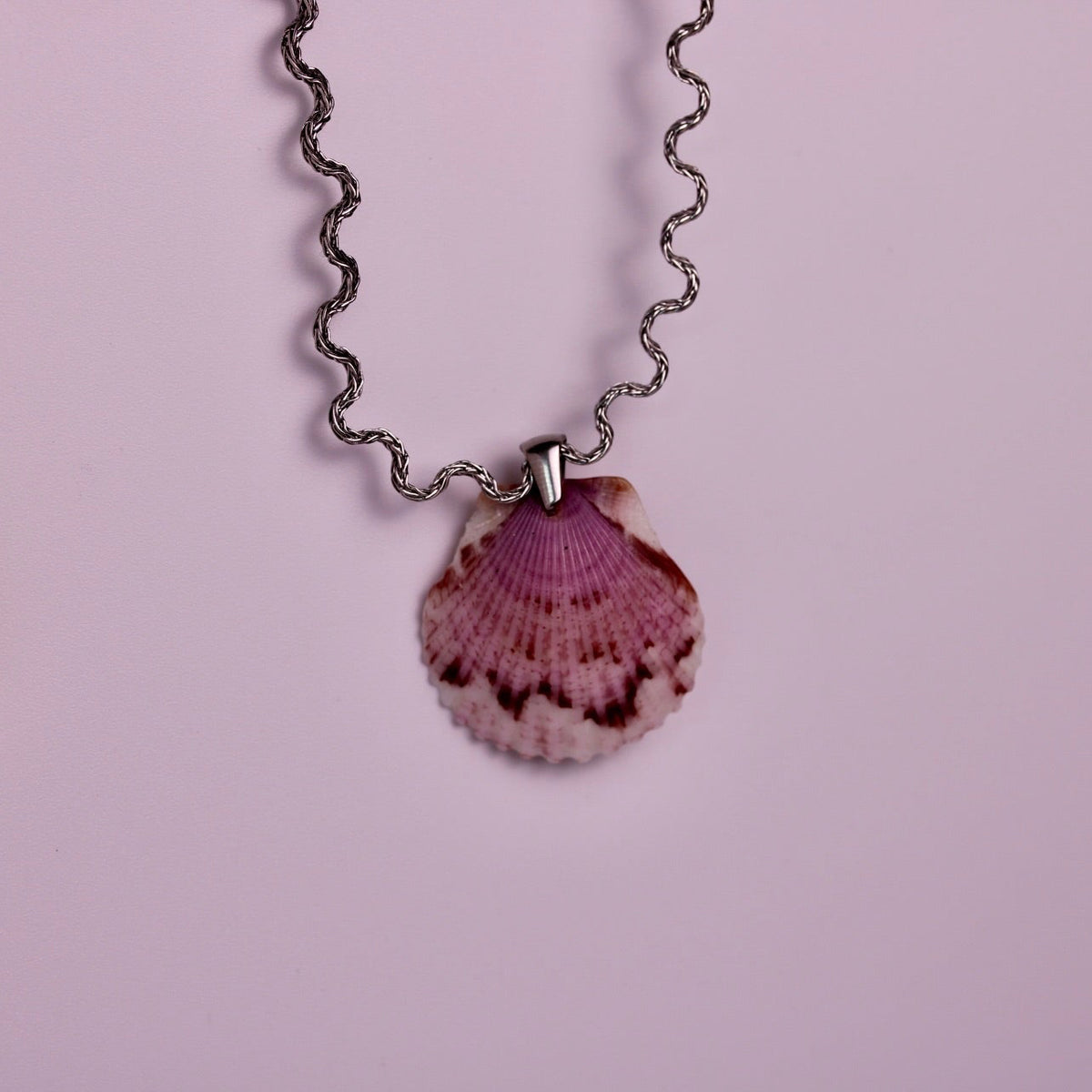 Two scallop shell pendant necklaces on silver chains, displayed against a solid light pink background. One necklace features a bright orange shell, and the other features a pink and white shell.