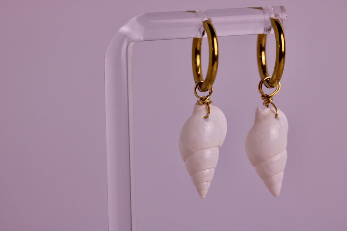 A pair of gold hoop earrings with white spiral seashell charms, hanging from a clear acrylic display stand against a solid lavender background.