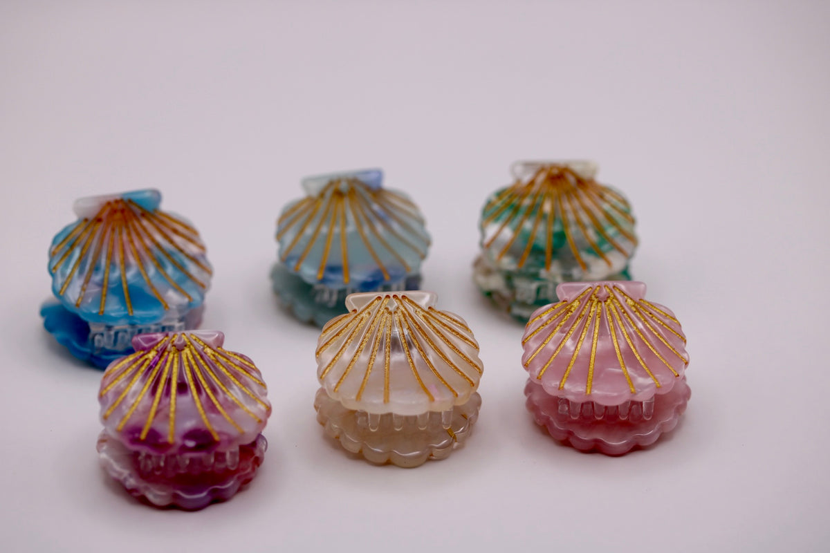 Six small shell-shaped decorative items arranged on a plain light background, each with a scalloped base and radiating ribbed top. The shells are translucent and come in blue, aqua, green, purple, beige, and pink, with metallic gold line detailing on top.