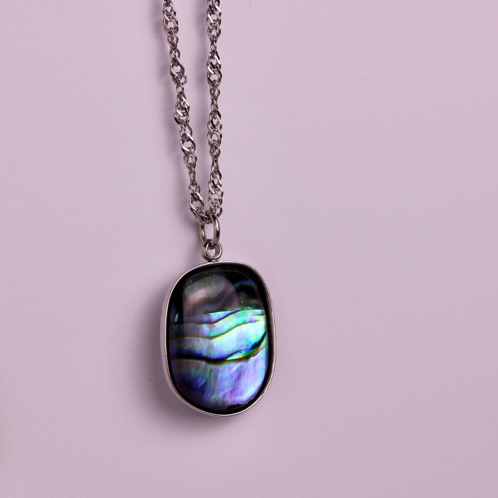 Two oval abalone shell pendant necklaces hanging against a soft pink background, one with a gold chain and one with a silver chain.