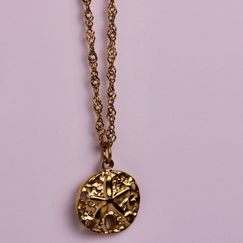 Two round pendant necklaces with textured starburst designs, one in silver and one in gold, displayed against a solid light pink background.
