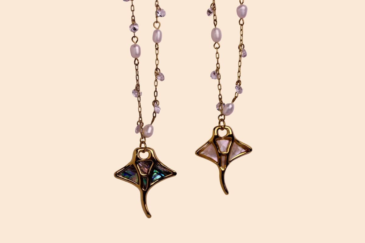 Two gold-toned beaded necklaces with mother-of-pearl pearl-like beads and iridescent abalone shell pendant charms, displayed against a solid light pink background.