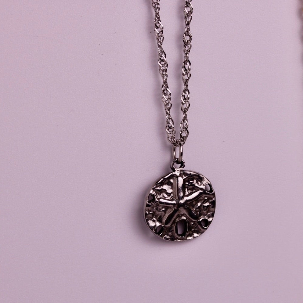 Two round pendant necklaces with Celtic-style knotwork designs, one silver-toned and one gold-toned, hanging against a solid light pink background.