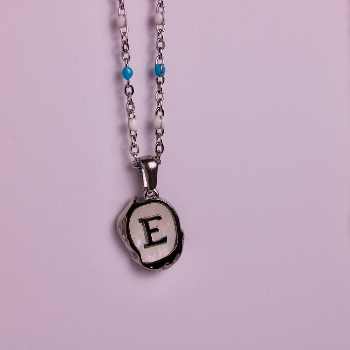 Two personalized initial necklaces hanging against a light pink background. The left necklace has a silver-tone chain with small blue and white beads and a silver pendant with a black-and-white shell-like border enclosing the letter 'E'.