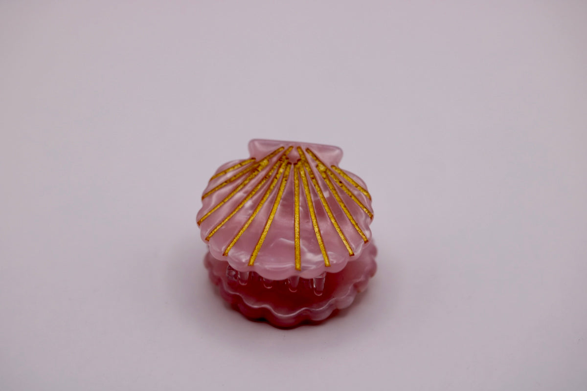 A small clam-shell-shaped decorative object with a translucent pink top, deep red base, and radiating metallic gold lines, centered on a plain light background.