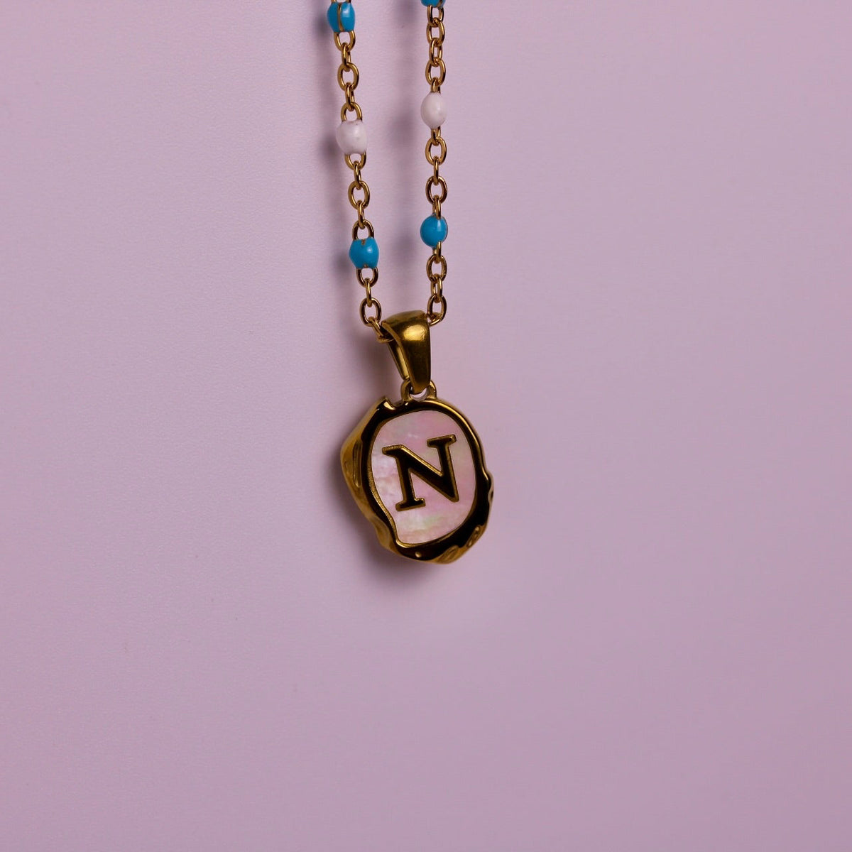 A gold initial necklace with a letter 'N' pendant on a light pink background, with another necklace partially visible on the left showing a letter 'E'.