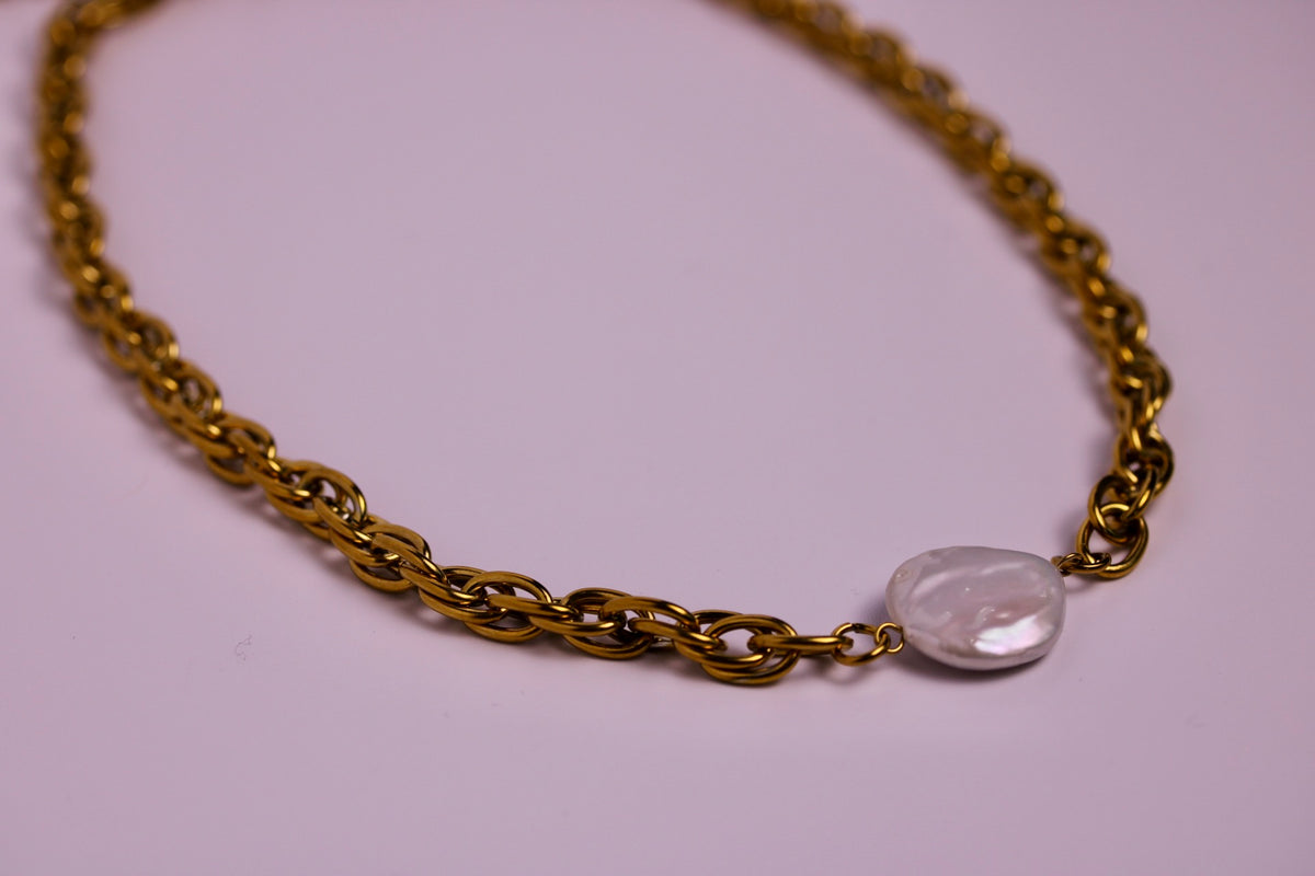 A gold chain necklace with a small, irregular, polished light gray stone pendant, displayed on a solid light pink background.