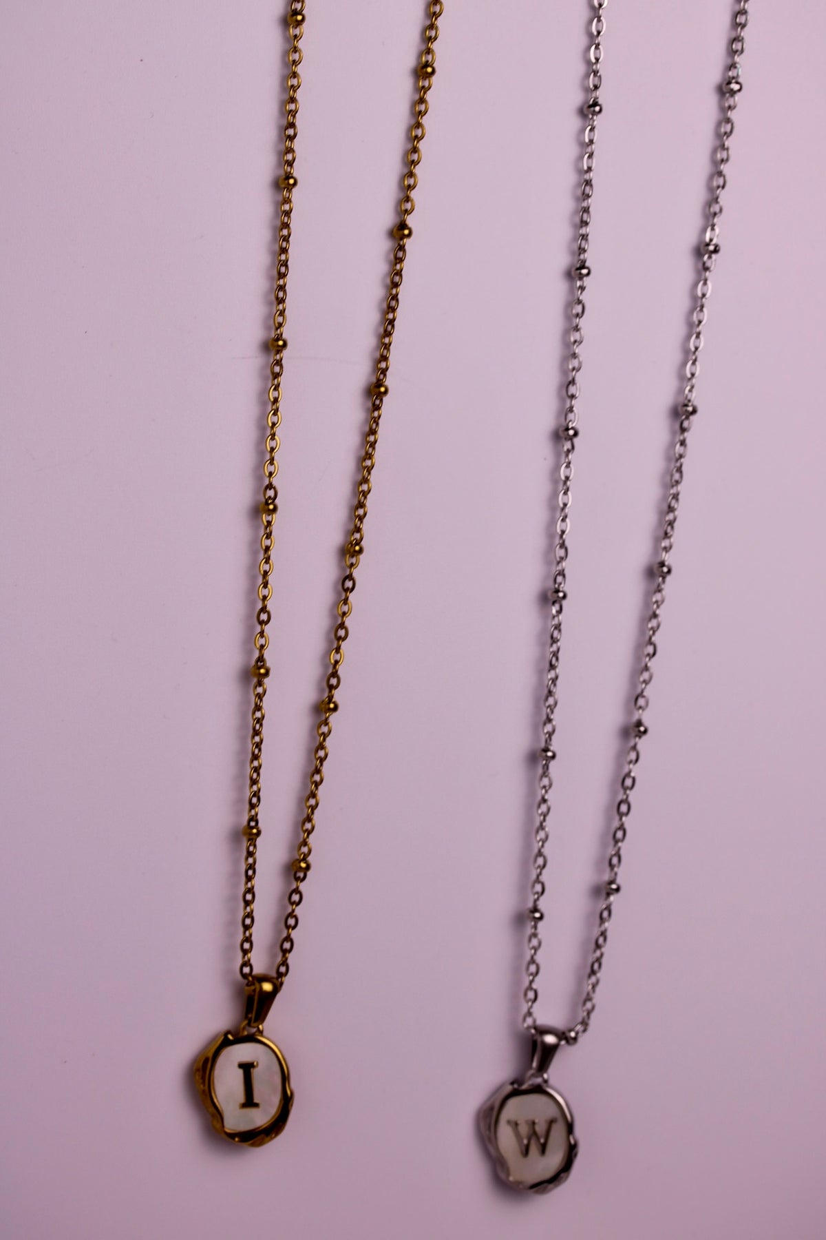 Two delicate necklace chains hanging against a light pink background. The left chain is gold-toned with a small oval pendant featuring the letter 'I' in cream on a dark background.
