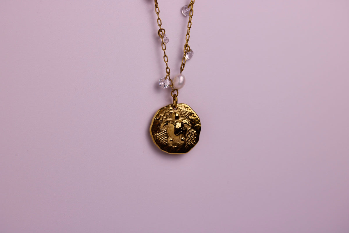 A gold coin pendant necklace with a textured circular medallion, hanging from a gold chain with small clear crystal and pearl accents against a solid light pink background.