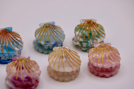 Five small shell-shaped decorative boxes or containers in pastel colors with gold radial lines, arranged on a plain light background.